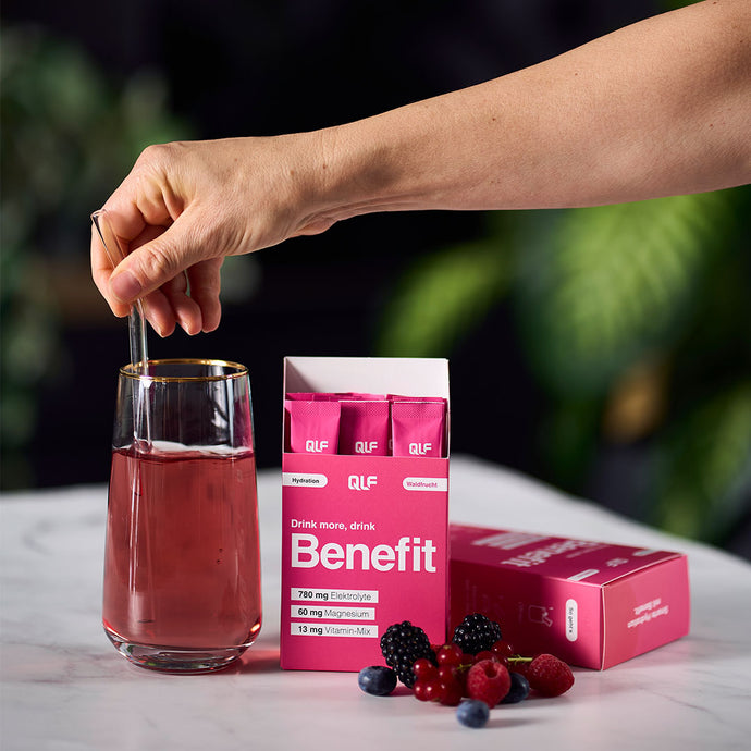 Hand stirring pink hydration drink in glass next to open pink Benefit box and fresh berries on marble surface