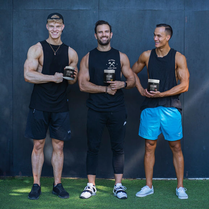 Drei Männer in Sportkleidung stehen im Freien nebeneinander, lächeln und halten schwarze Behälter mit der Aufschrift "VOLUME" in der Hand. Sie stehen vor einer dunklen Wand auf einer Wiese.