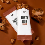 Tasty Bar Proteinriegel Brownie in Verpackung und aufgebrochen, umgeben von Schokostücken auf braunem Untergrund, Galerieposition 1