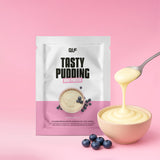 Eine Packung High Protein Tasty Pudding steht vor einem rosafarbenen Hintergrund neben einer Schüssel mit Pudding. Ein Löffel hebt die proteinreiche Leckerei an, in der Nähe liegen frische Blaubeeren, die den Muskelaufbau unterstützen.