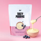Eine Packung High Protein Tasty Pudding mit einer Schale Pudding, Blaubeeren und einem Löffel, der den Pudding ausgießt. Der rosa Hintergrund enthält 19 g Protein, 87 kcal und 1,9 g Kohlenhydrate pro Portion - ideal für den Muskelaufbau.