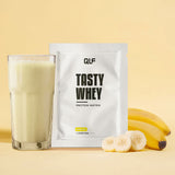 Tasty Whey Proteinpulver Banane mit Shake im Glas, Bananenscheiben und Bananen im hellen Hintergrund Galerie 1