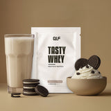 Tasty Whey Proteinpulver Cookies & Cream Sachet mit Shake im Glas, Keksen und Sahnecreme vor beige-braunem Hintergrund