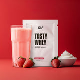 Tasty Whey Erdbeer Sahne Proteinpulver Sachet mit cremigem Shake im Glas, frischen Erdbeeren und Sahne vor rotem Hintergrund