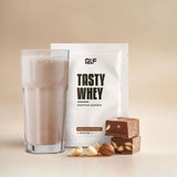 Tasty Whey Proteinpulver Haselnuss Nougat mit cremigem Shake im Glas und Nougatstücken vor hellem Hintergrund
