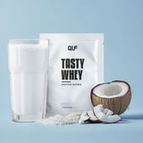 Tasty Whey Proteinpulver Kokos mit Shake im Glas, Kokosraspeln und frischer Kokosnuss vor hellem blauem Hintergrund