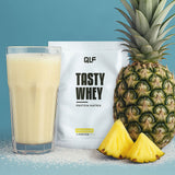 Tasty Whey Pina Colada Proteinpulver Sachet mit cremigem Shake im Glas und frischer Ananas vor blauem Hintergrund