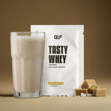 Tasty Whey Salted Caramel Proteinpulver Sachet mit Shake im Glas und Karamellstücken vor braunem Hintergrund