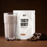 Tasty Whey Schokolade Proteinpulver Sachet mit Schokoshake im Glas und Schokostücken vor braunem Hintergrund Galerie 1