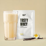 Tasty Whey Vanille Proteinpulver Sachet mit cremigem Shake im Glas, Vanilleschote und Blüte vor beigem Hintergrund