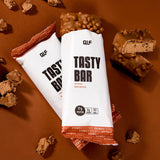 Tasty Bar Proteinriegel Brownie in Verpackung und aufgebrochen, umgeben von Schokostücken auf braunem Untergrund, Galerieposition 1