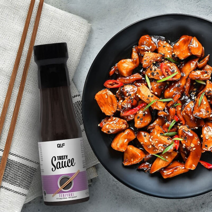 A black bowl filled with glazed chicken pieces garnished with chili slices and sesame seeds next to a bottle of QLF Teriyaki sauce.