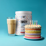 Tasty Whey Proteinpulver Birthday Cake mit cremigem Shake im Glas und bunter Geburtstagstorte vor blauem Hintergrund