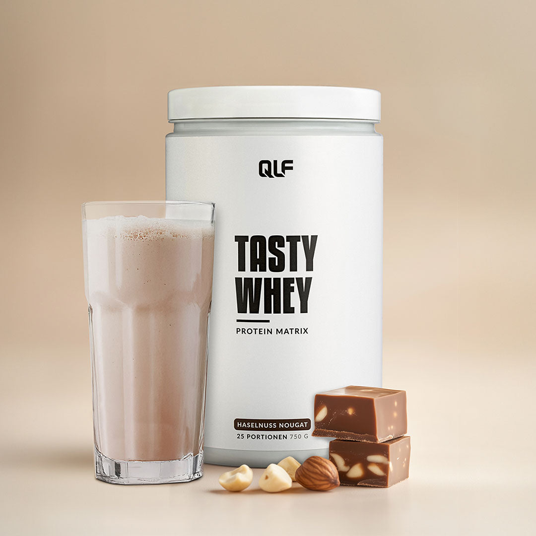 Tasty Whey Proteinpulver Haselnuss Nougat mit Glas Shake, Nüssen und Schokostücken auf beige Hintergrund, Galerie 1