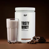 Tasty Whey Proteinpulver Schokolade weniger süß mit cremigem Shake im Glas und Schokostücken vor braunem Hintergrund