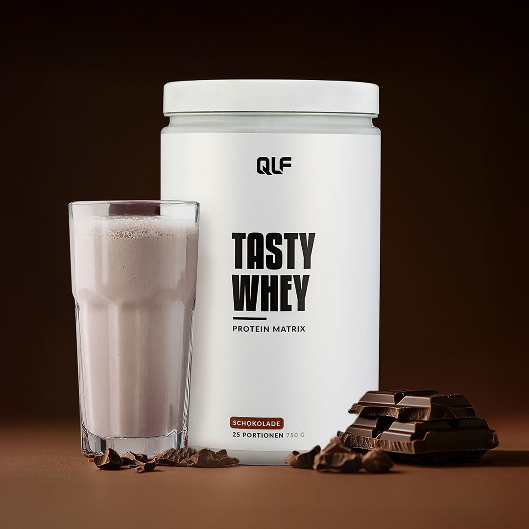 Tasty Whey Proteinpulver Schokolade mit cremigem Shake im Glas und Schokostücken vor braunem Hintergrund Galerieposition 1