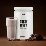 Tasty Whey Proteinpulver Schokolade mit cremigem Shake im Glas und Schokostücken vor braunem Hintergrund Galerieposition 1