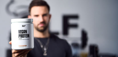 Man holding white container labeled Vegan Protein in front of blurred background with QLF logo on wall